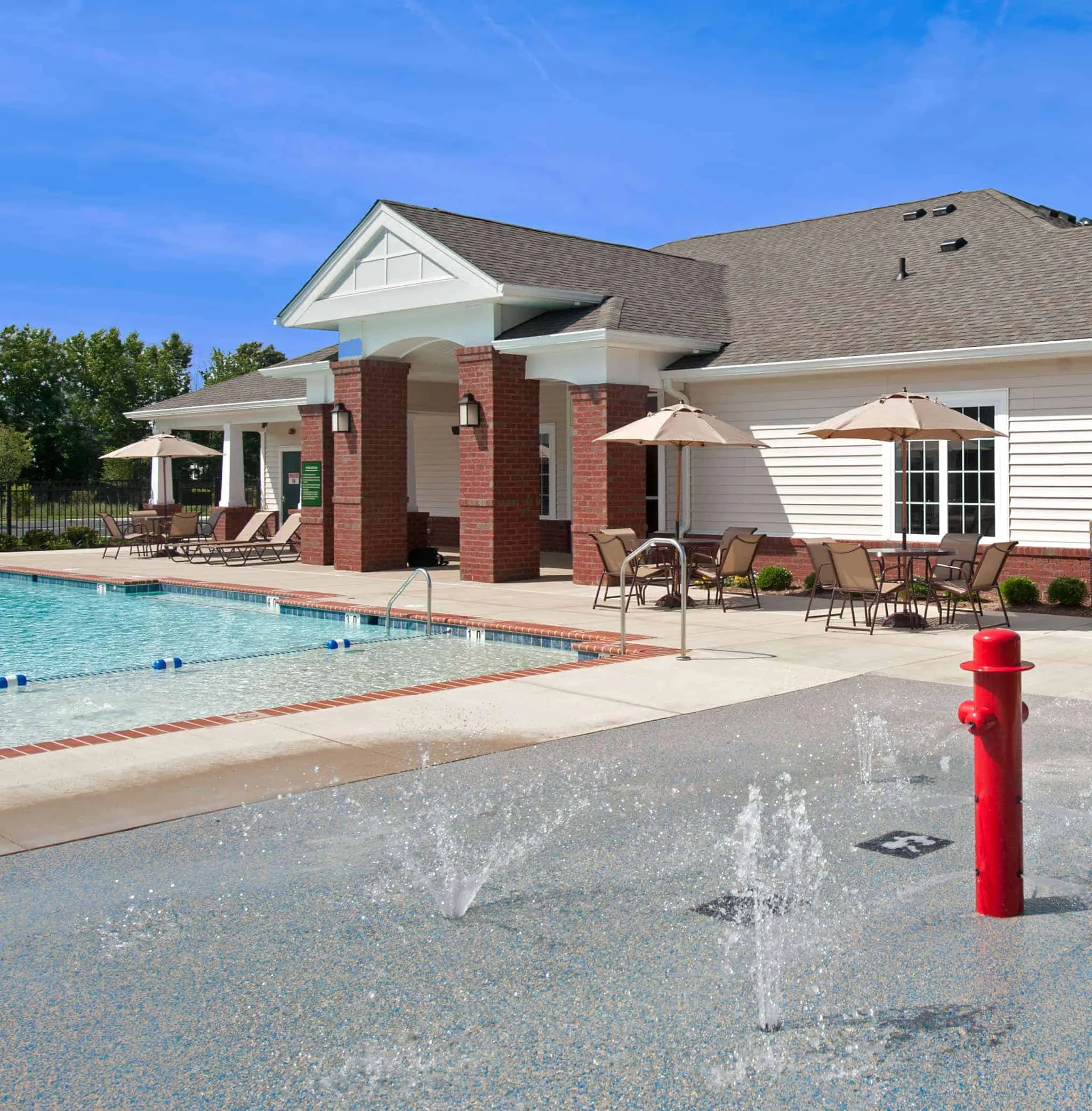An outdoor children's splash pad area featuring water jets and a red fire hydrant sprayer next to the community swimming pool and clubhouse